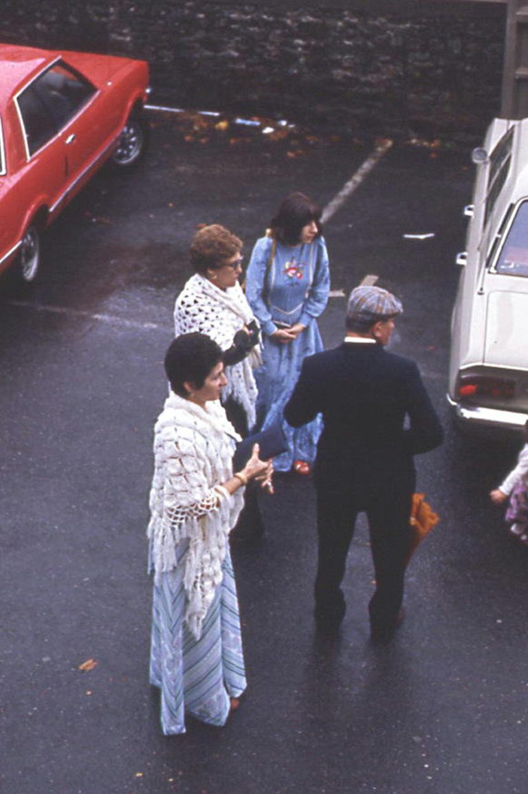 Adults gather in a vintage parking lot conversation, dressed in classic fashion.