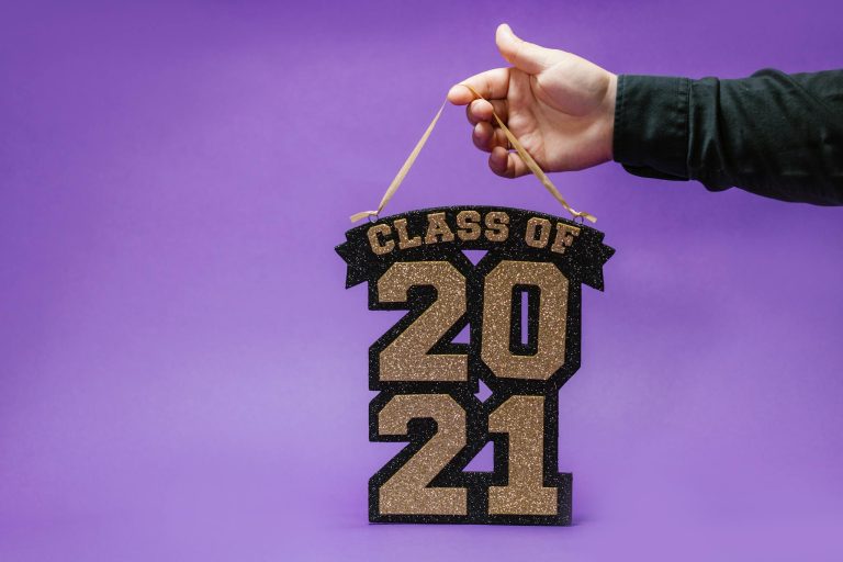 Glittery 'Class of 2021' sign held by a hand against a purple background.